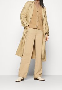 Beige trenssitakki neulotun ruskean neuleen päällä, yhdistettynä beigen värisiin leveälahkeisiin housuihin ja kermaisiin nilkkabootseihin, joissa on nappi- ja taskuyksityiskohtia.