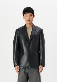 Schwarze Lederblazer mit eingeknöpften Revers, einreihiger Front, zwei Taschen und dezente Nähdetails. Getragen über einem beigen Hemd.