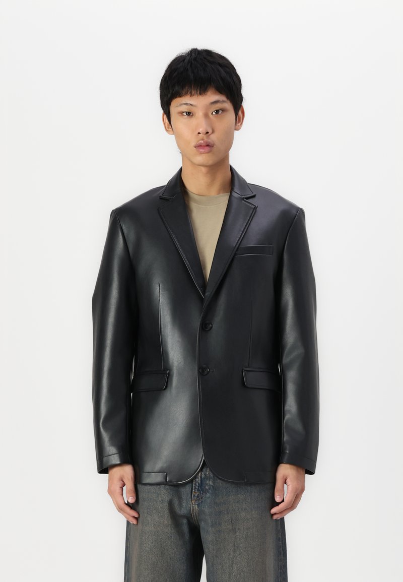 Schwarze Lederblazer mit eingeknöpften Revers, einreihiger Front, zwei Taschen und dezente Nähdetails. Getragen über einem beigen Hemd.