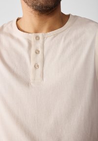 Beige puuvillainen henley-paita, jossa on pyöreä kaula-aukko, kolme napin aukkoa kirkkailla napeilla ja pehmeä, sileä tekstuuri.