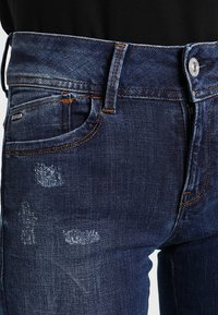 Vaqueros de mezclilla azul oscuro con ligeros desgastes, botón plateado, costuras naranjas y diseño tradicional de cinco bolsillos. Textura suave.