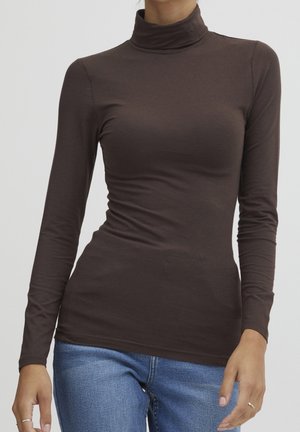 Personne portant un haut à col roulé moulant à manches longues marron foncé et un jean bleu clair, debout devant un fond clair uni.