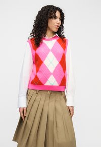 Πλεκτό γιλέκο χωρίς μανίκια σε ροζ, κόκκινο και εκρού αργκλάι μοτίβο, συνδυασμένο με λευκό πουκάμισο με γιακά και χακί πλισέ φούστα. Μαλακή υφή.