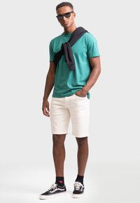 Grünes Baumwoll-T-Shirt, weiße Denim-Shorts, schwarzer Pullover über die Schultern drapiert, schwarze Sonnenbrille und schwarze Sneakers mit weißen Akzenten.