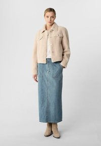 Giacca beige in finta pelliccia shearling con due tasche sul petto, abbinata a una lunga gonna in denim e stivaletti alla caviglia in tonalità neutre. Tessuti lisci e texture.