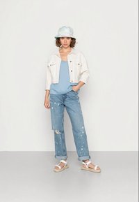 Jaqueta de ganga branca, t-shirt sem mangas azul-claro, jeans claros desgastados com manchas de tinta e sandálias brancas de tiras, complementados por um chapéu de balde em tons pastéis.