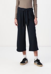 Navy wide-leg trousers with a tie waistband, made from smooth fabric. Styled with a ribbed beige top and black sneakers.