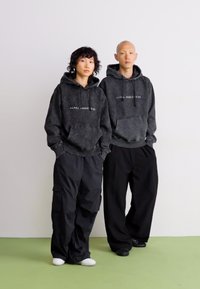 Du žmonės vilkintys tamsiai pilkas pūkuotas maikes su užrašu "ALPHA INDUSTRIES", juodas laisvas kelnes, stovintys šalia vienas kito prieš neutralų foną.