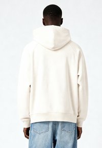 Sweat à capuche couleur crème avec capuche à cordon, manches raglan et ourlet côtelé. Le tissu semble doux et lisse, associé à un jean en denim bleu.