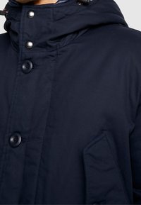 Granatowa kurtka z kapturem o gładkiej teksturze, z dużymi czarnymi guzikami, podniesionym kołnierzem oraz detalem w postaci bocznej kieszeni.