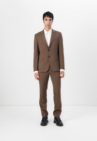 Costum maro, croit cu o jachetă ajustată și pantaloni drepți. Textură netedă, design cu un singur rând de nasturi și pantofi negri. Cămașă albă cu nasturi dedesubt.