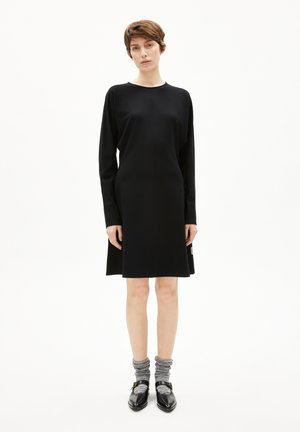 Schwarzes Langarmkleid mit lockerer Passform, runder Ausschnitt und leicht ausgestellter Form, kombiniert mit grauen strukturierten Socken und schwarzen Schuhen.
