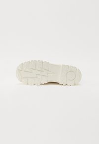 Suela de zapatilla de goma blanca con patrón en zigzag texturizado, que presenta tacos triangulares para agarre y un logo de marca circular en el talón.