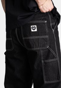 Pantaloni in denim neri con cuciture bianche, multiple tasche e un'etichetta quadrata del marchio. Le cuciture a vista aggiungono texture e dettaglio.