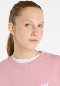 Sweat-shirt rose avec un col côtelé et un logo visible. Texture de tissu lisse, présentant une coupe décontractée et un design simple.