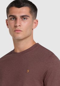 T-shirt in cotone marrone con scollatura rotonda e maniche corte. Presenta un piccolo logo ricamato giallo sul lato sinistro del petto.