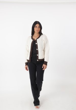 Cárdigan de ganchillo en crema y marrón oscuro, con cuello redondo, botones en la parte delantera y mangas largas, combinado con pantalones negros. Textura suave.
