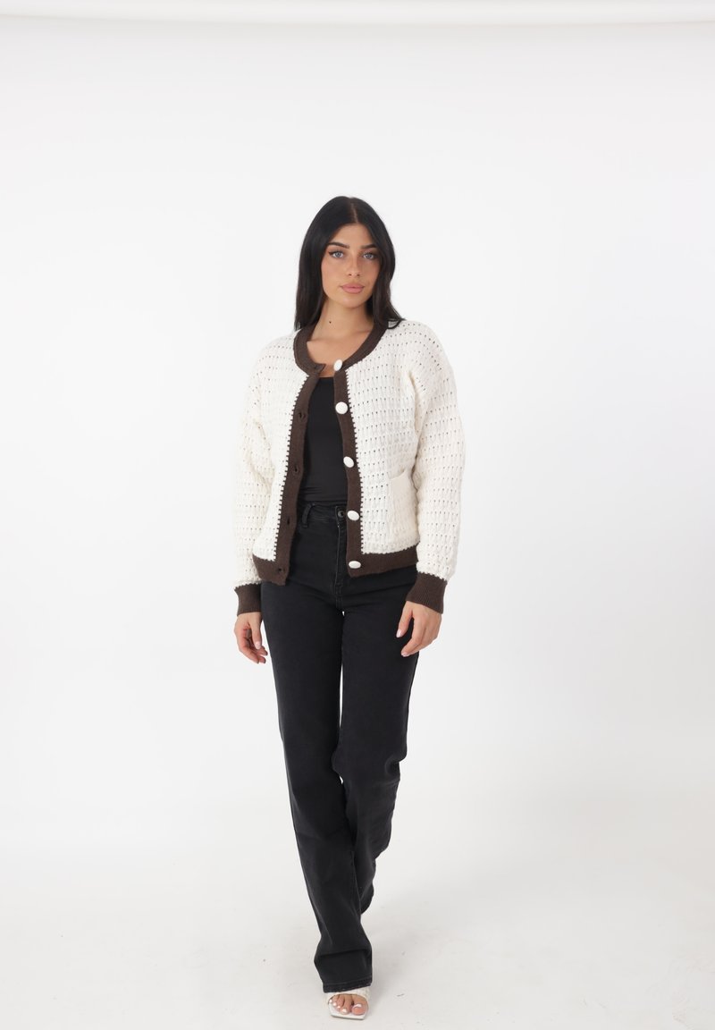 Cárdigan de ganchillo en crema y marrón oscuro, con cuello redondo, botones en la parte delantera y mangas largas, combinado con pantalones negros. Textura suave.