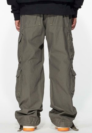 Vue de dos d'une personne portant un pantalon cargo ample vert olive avec plusieurs poches et des chaussures à semelle beige sur un fond uni.