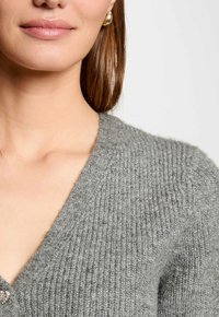 Grijze gebreide cardigan met een V-hals en een gestructureerd patroon, met subtiele glans. Verdere details zijn een kleine versierde knop.