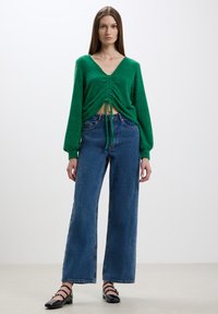 Maglione verde, soffice e corto con scollatura a V e dettaglio davanti arricciato, abbinato a jeans a gamba larga in denim blu scuro e scarpe nere flat.
