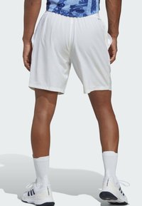 Hvide atletiske shorts med elastisk talje, sidelommer og en glat tekstur, båret med hvide sokker og sko med sorte detaljer.