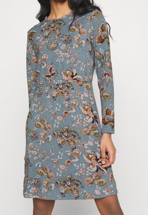 Frau trägt ein blaues Kleid mit langen Ärmeln und mehrfarbigen Blumen- und Schmetterlingsmustern, steht vor einem einfarbigen Hintergrund.