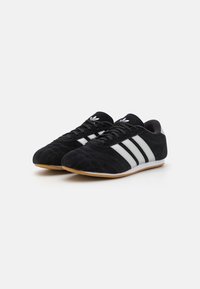 Black suede sneakers with three white stripes, flat gum sole, rounded toe, and lace-up design. Textured upper with stitching details.
