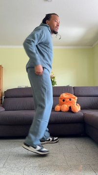 Fato de treino azul claro com capuz, usado com ténis pretos e brancos. Polvo de pelúcia cor-de-laranja brilhante sobre um sofá castanho escuro. Fundo de parede verde.