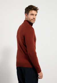 Kupferfarbener Strickpullover mit hohem Kragen, taillierter Passform und gerippten Bündchen. Getragen mit dunkelblauen Cordhosen.