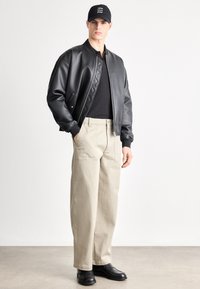 Fekete bőr bombázó dzseki, könnyű fekete póló, bézs bő szárú nadrág, fekete bokacsizma, és fekete sapka fehér felirattal.