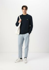 Maglione a maniche lunghe blu navy con un piccolo logo giallo, pantaloni tapered azzurri chiari, scarpe da ginnastica bianche e una borsa tote beige. Texture del tessuto lisce.