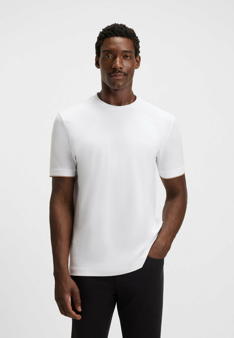 Witte kraagloze t-shirt met korte mouwen, gladde textuur en een lichte glans, gecombineerd met zwarte broek. Minimalistische branding zichtbaar.