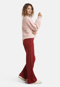 Pink knitted sweater with a textured pattern, paired with red wide-leg pants and white sneakers. Model stands sideways, showcasing outfit.