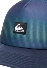 Modrá gradientová čepice z texturované tkaniny, s vodorovnými pruhy. Na přední straně má černou nášivku loga Quiksilver.