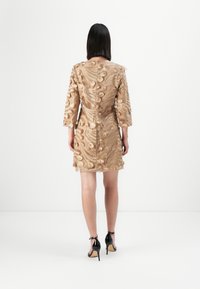 Beige brodert kjole med teksturerte blomster mønstre, trekvart ermer og en tett silhuett, parret med svarte høye hæler.