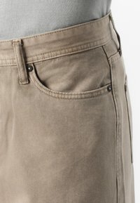 Close-up of a pair of light tan pants, showing the waistband and pocket detail, with a grey shirt partially visible above.