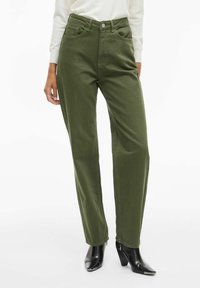 Pantalones de tiro alto de color verde oliva con un diseño de pernera recta, con bolsillos frontales y cierre de botón, complementados con botas de tobillo de tacón negro.