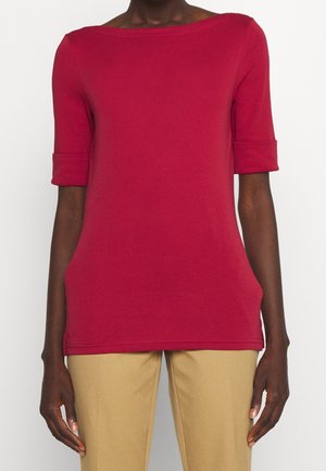 Persona che indossa una maglietta rossa a maniche corte con polsini risvoltati e pantaloni beige, mostrata dal collo ai fianchi su uno sfondo semplice.