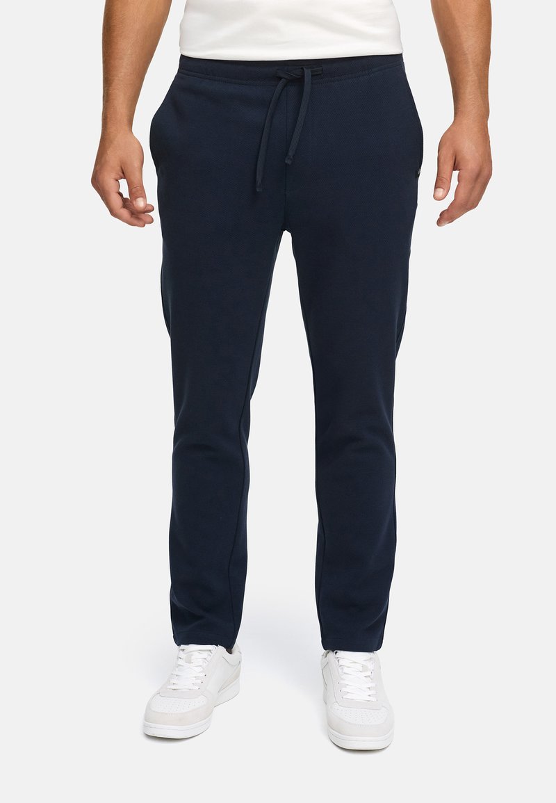 Mand iført navy blå slim-fit sweatpants med snor og hvide sneakers, stående foran en ensfarvet baggrund.