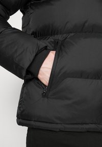 Main dans la poche zippée d'une veste d'hiver noire matelassée avec un poignet côtelé, montrée de la taille au coude.