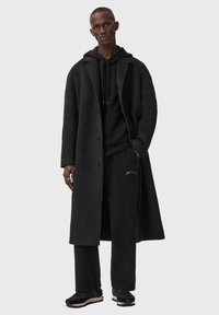 Zwarte wollen overjas met grote revers en knopen, gedragen over een zwarte hoodie en joggingbroek, gecombineerd met zwarte sportschoenen.