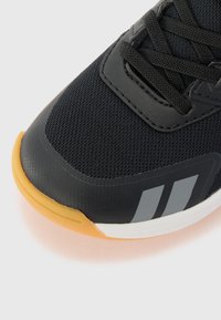 Chaussure de sport noire avec une tige en maille texturée, semelle en caoutchouc et rayures grises. Présente des lacets noirs et un capuchon de orteil beige.