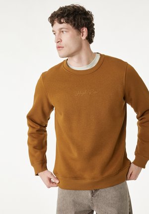 Giovane uomo che guarda a sinistra, indossa una felpa marrone con la scritta "High Rise" e pantaloni grigio chiaro su uno sfondo bianco uniforme.