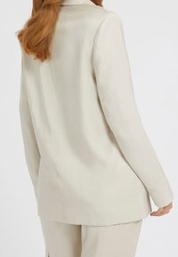 Vaaleanbeige bleiseri, jossa on sileä, hieman kiiltävä pinta, pitkät hihat ja jämäkkä muotoilu, nähtynä takaa.