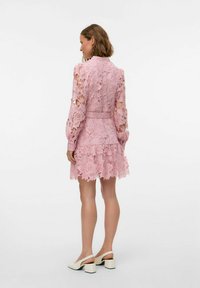 Robe en dentelle rose à manches longues et motif floral, présentant un corsage ajusté, une jupe à volants et une ceinture assortie. Portée avec des chaussures à talons carrés blanches.