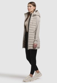 Beige puffer vest med hætte, udstyret med quiltede paneler og sidelommer, parret med sorte leggings og lyse sneakers.