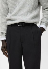 Uomo che indossa un maglione a costine grigio chiaro infilato in pantaloni neri a vita alta con una cintura in pelle nera, con una mano in tasca.