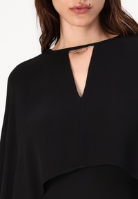 Črna bluzica z V-izrezom in akcentom iz srebrne verige, izdelana iz gladkega materiala z udobnim krojem in plastenjem.