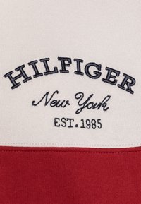 Felpa con girocollo caratterizzata da una parte superiore beige con la scritta ricamata "HILFIGER" e "New York" in blu navy, e una parte inferiore rossa.